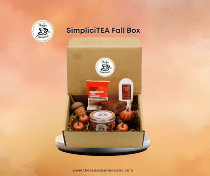 SimpliciTEA Fall Box gift set displayed in an open kraft cardboard box, featuring a chocolate bar, tea tin, candle, acorns, and small pumpkins, set against a warm orange gradient background.